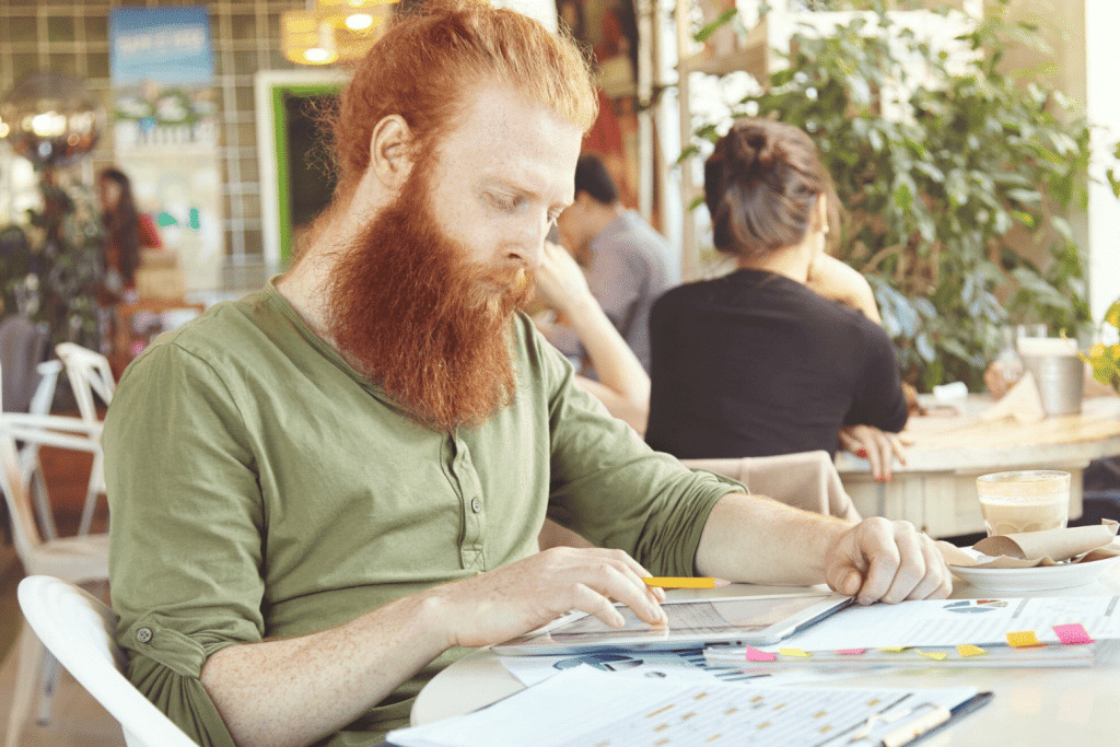 A man with a long red beard and green shirt sits at a table in a café, looking at documents and holding a pencil. Papers and a glass of coffee are on the table; other people are seated in the background.